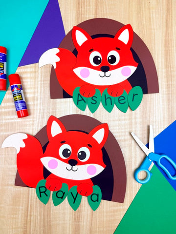 Two paper fox crafts with names 'Asher' and 'Ray' on a wooden surface with glue and scissors.