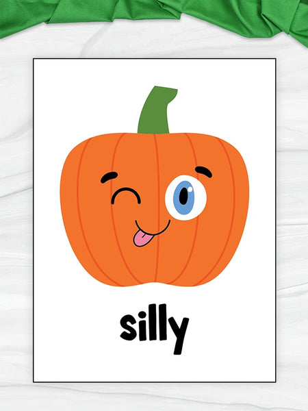 A flashcard featuring a cute cartoon pumpkin with a winking blue eye and the word 'silly' in black lowercase text below it, displayed on a light wood surface with a green fabric accent at the top.