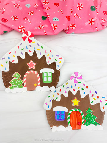 Two colorful gingerbread house craft on a white surface with a pink fabric background.