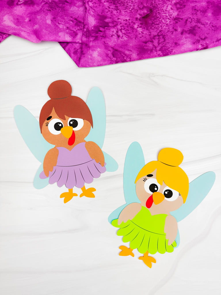 Two paper craft fairy turkey in disguises on a white background with a purple fabric in the corner.