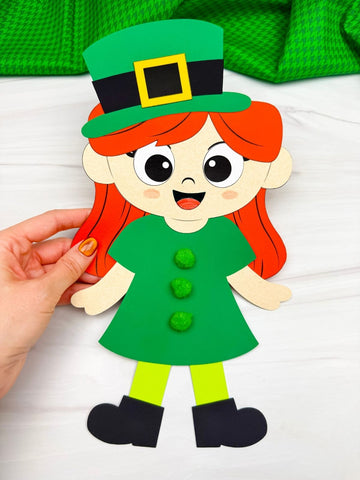 Hand holding a paper cutout of a girl leprechaun with red hair, green hat, and outfit on a white background.