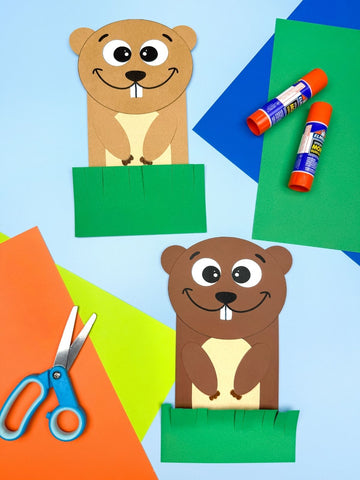 Two paper crafts of groundhogs on a colorful background with scissors and glue sticks.
