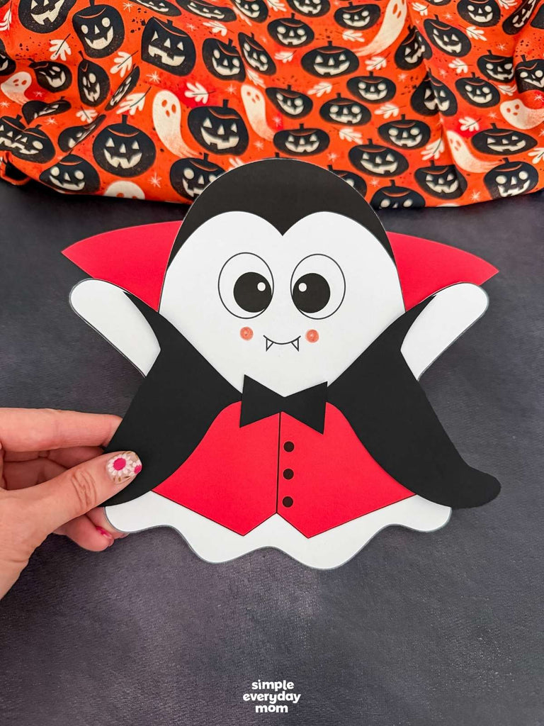 A hand holds a paper-cutout vampire ghost disguise ghost in front of an orange Halloween pumpkin fabric. The craft is white with a black face and hair, a red cape and vest, and a black bow tie.