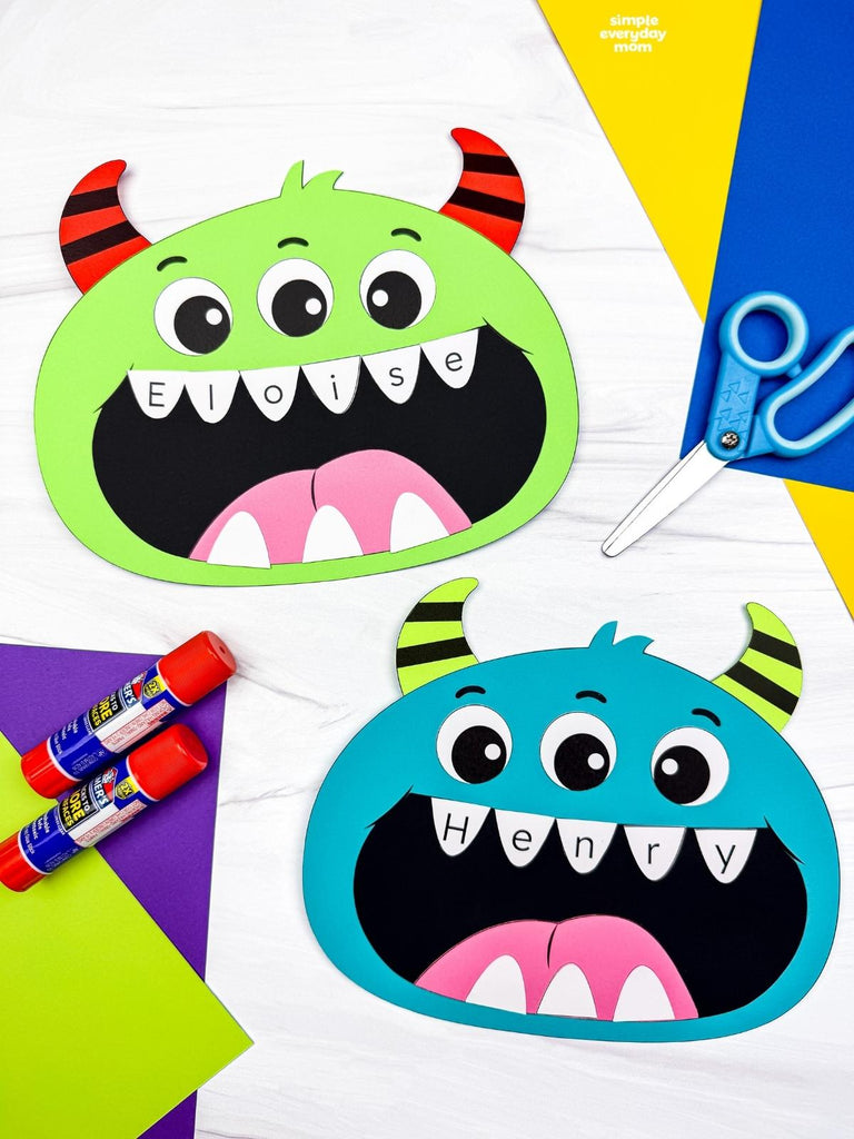 Two colorful monster face craft cutouts on a white background, one green with red horns and one blue with yellow horns, surrounded by craft supplies including glue sticks, scissors, and colored paper.