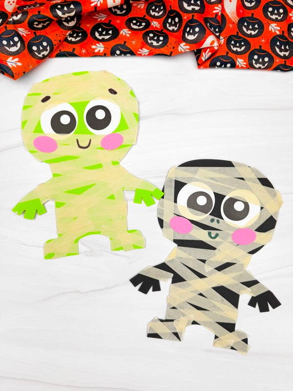 Two Halloween mummy paper crafts on white background with orange Halloween fabric on top