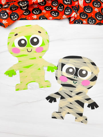 Two Halloween mummy paper crafts on white background with orange Halloween fabric on top
