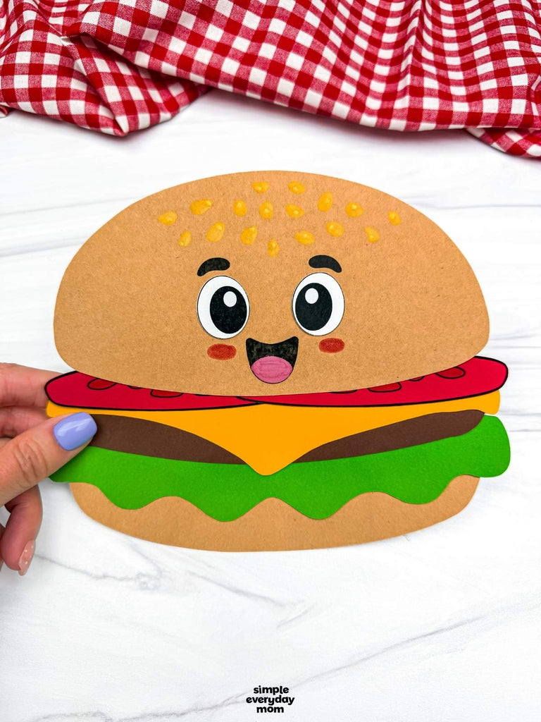 Paper craft of a cartoon hamburger held by a hand on a white background with a red and white checkered fabric.