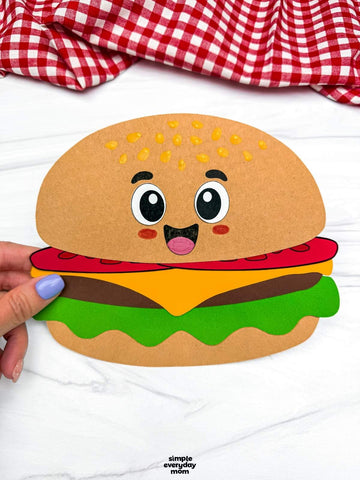 Paper craft of a cartoon hamburger held by a hand on a white background with a red and white checkered fabric.