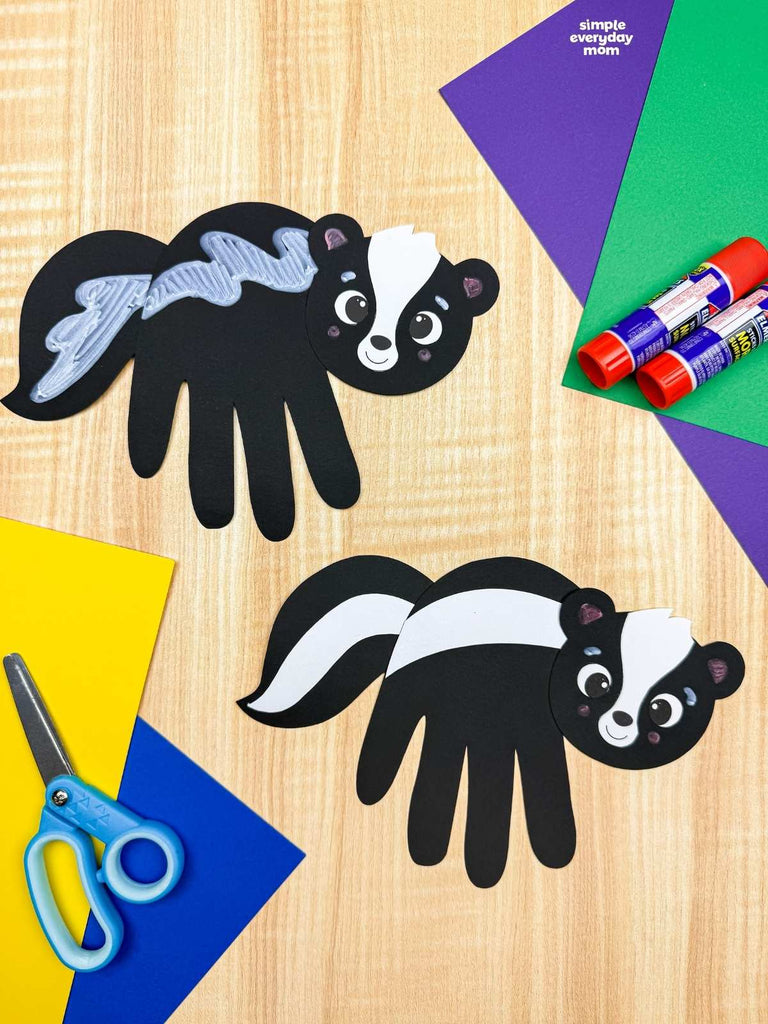 Two black and white skunk handprint crafts displayed on a light wood background with craft supplies including colored paper, scissors, and glue sticks.