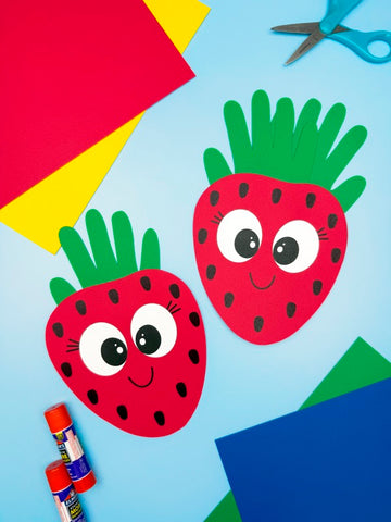 2 handprint strawberry crafts on a blue background with colorful paper, scissors, and glue sticks in the background