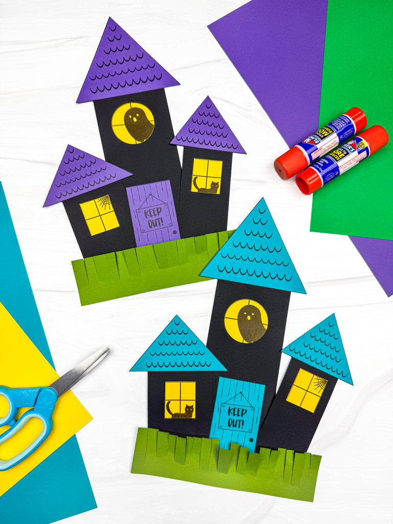 Two colorful paper haunted house crafts made from black, blue, purple, green, and yellow cardstock, displayed with scissors, glue sticks, and craft paper.