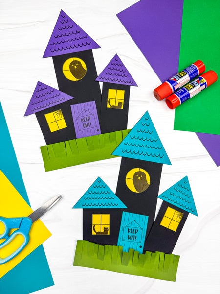 Two colorful paper haunted house crafts made from black, blue, purple, green, and yellow cardstock, displayed with scissors, glue sticks, and craft paper.