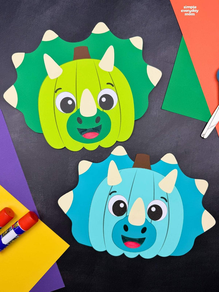 Two colorful paper crafts shaped like triceratops pumpkins, one green and one blue, displayed with construction paper, glue sticks, and scissors on a dark background.