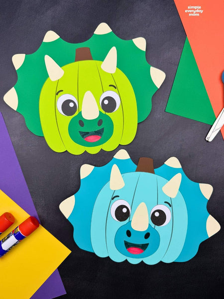 Two colorful paper crafts shaped like triceratops pumpkins, one green and one blue, displayed with construction paper, glue sticks, and scissors on a dark background.