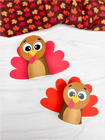 Two paper cup turkey crafts on a white surface with a patterned fabric background.