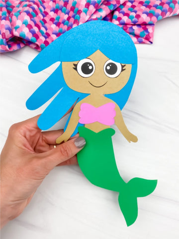 Handmade mermaid craft with blue hair and green tail held by a hand on a white background.