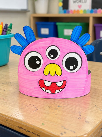 A pink monster head craft hat with blue spiky ears, large cartoon eyes, a yellow nose, and a red smile with white teeth, sitting on a wooden desk in a classroom.
