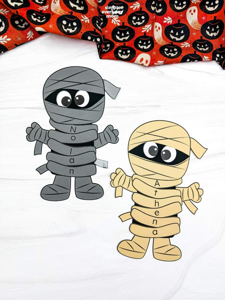 Two mummy name crafts on a white surface with Halloween fabric on top 