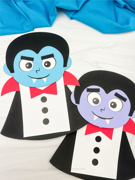 Two paper vampire crafts with blue and purple faces, black hair and capes, red collars, white shirts, buttons, and small fangs are displayed on a white surface.