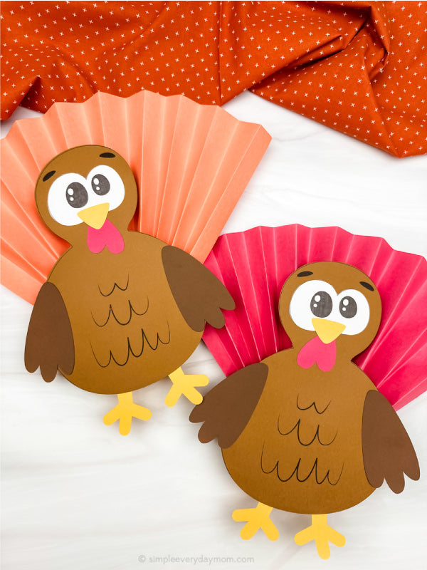 Two paper turkey cutouts on a white background with orange and pink cupcake accordion fold papers.