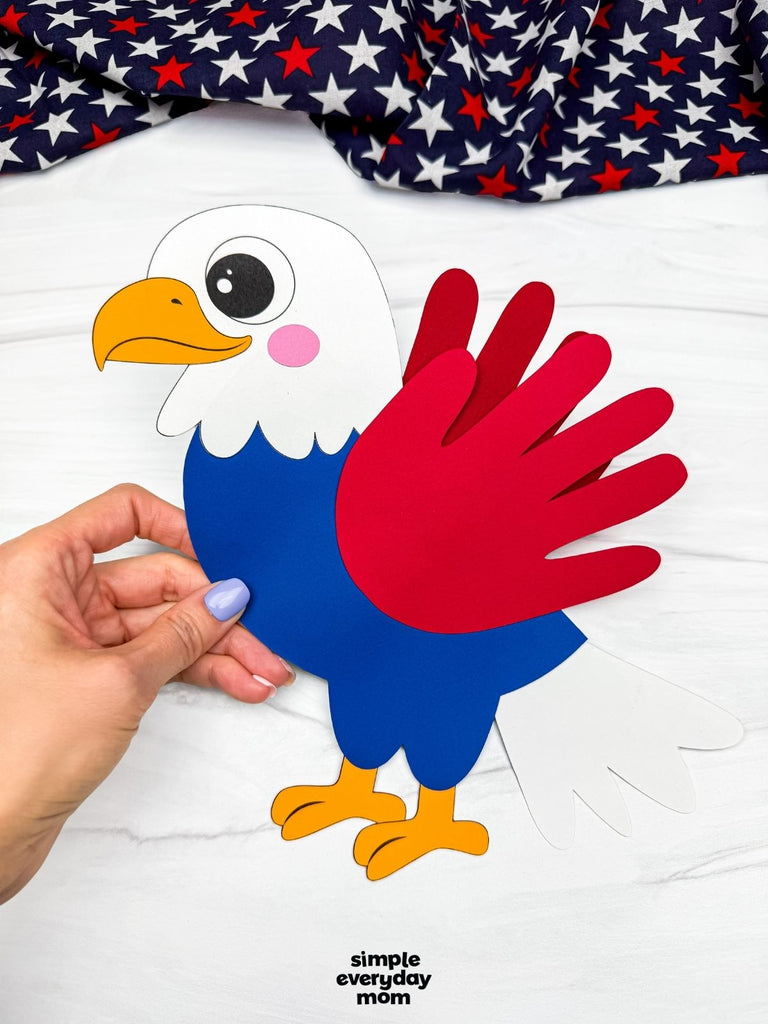 Handmade paper eagle craft held by a hand with a red, white, and blue fabric on top.