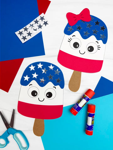 2 finished patriotic popsicle crafts for kids with scissors, glue sticks, star stickers and colored paper in the background