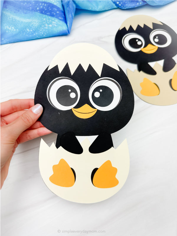 Hand holding a paper craft of a penguin hatching from an egg on a white background.