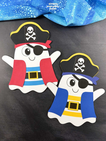 Two cute ghost-shaped pirate crafts on a dark gray background with a blue fabric at the top. One ghost wears a red vest and the other wears a blue vest, both with yellow and black pirate hats and eye patches.
