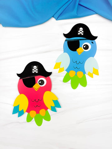 Two colorful paper parrot crafts, one red and one blue, each wearing a black pirate hat with a skull and crossbones and a black eye patch over one eye. The parrots have layered wings and feet in bright colors and are placed on a white surface with a blue cloth in the background.