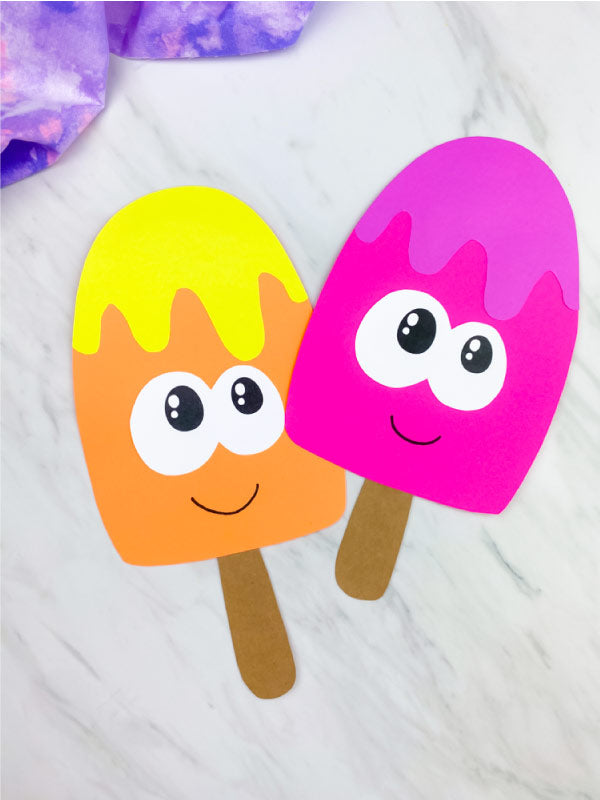 Two popsicle paper crafts on a white background with purple fabric on top 