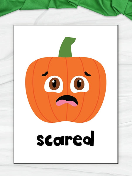 A flashcard featuring a cartoon orange pumpkin face with a green stem and the word 'scared' in black lowercase letters below. The print is photographed on a light surface with a green fabric strip at the top edge.