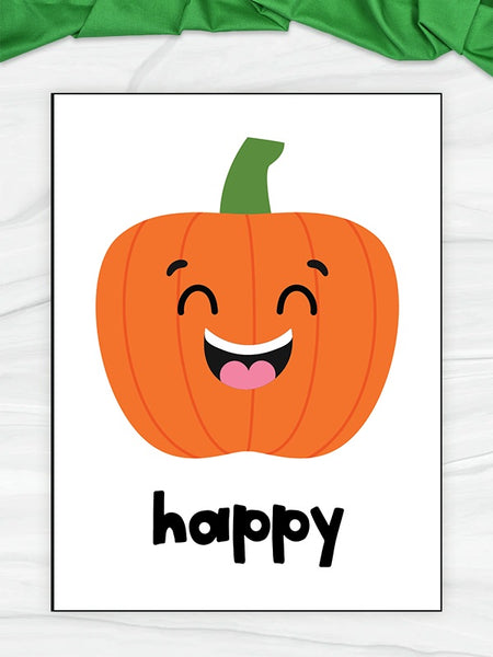 A flashcard featuring a smiling orange pumpkin with a green stem and the word 'happy' in black lowercase letters below. The print is photographed on a light surface with a green fabric strip at the top.
