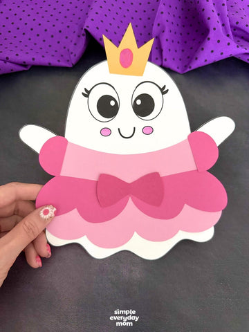 A cute ghost-shaped paper craft character wearing a gold crown and pink tutu dress, held by a hand against a dark gray background with purple polka-dot fabric in the upper corner.