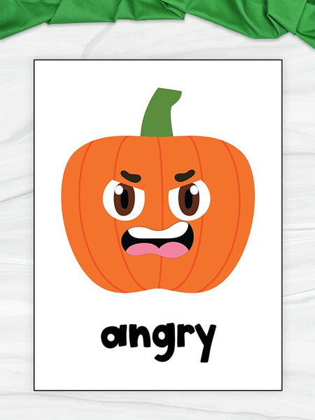 A children's flashcard featuring a cartoon angry pumpkin with the word 'angry' printed below it, displayed on a white marble surface with a green fabric accent at the top.