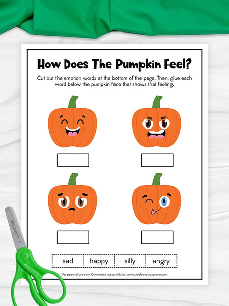 A printable pumpkin emotion worksheet titled "How Does The Pumpkin Feel?" shown on a white surface with a green fabric roll at the top and green-handled scissors at the bottom left. The worksheet features four cartoon pumpkins with different facial expressions and blank boxes underneath, plus emotion words at the bottom.