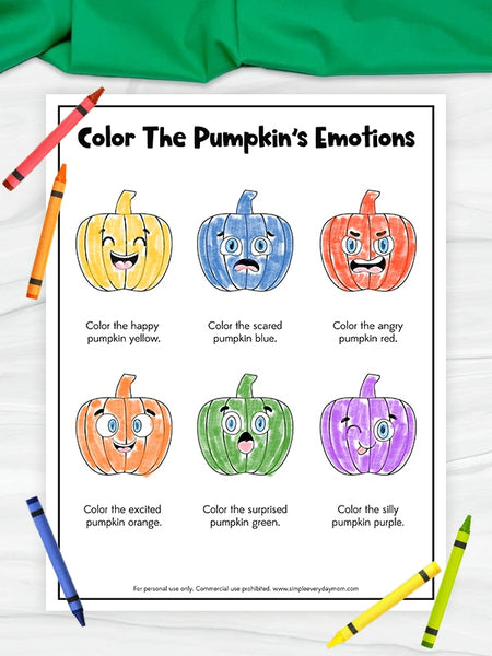 A printable coloring worksheet titled 'Color The Pumpkin's Emotions' showing six cartoon pumpkins in different colors with instructions to color them by emotion. The worksheet is displayed on a white wood background with crayons around it and a green fabric at the top.