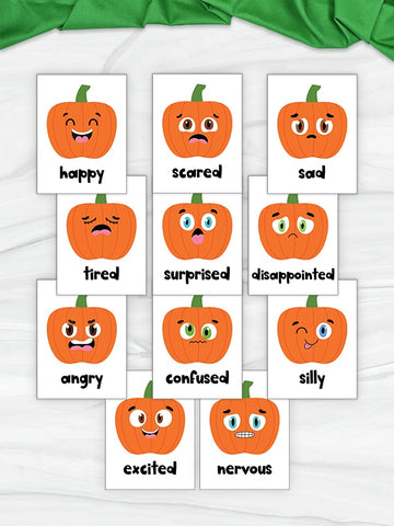 A set of white rectangular cards featuring cartoon orange pumpkins with different facial expressions and labels such as happy, scared, sad, tired, surprised, disappointed, angry, confused, silly, excited, and nervous, arranged on a light fabric background with a green fabric accent at the top.