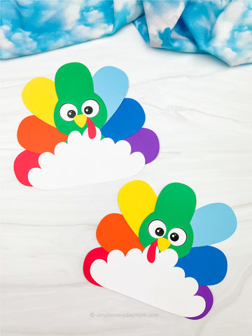 Rainbow paper turkey disguise craft on a white background with blue fabric in the corner.