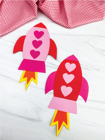 Two paper rockets with heart designs on a white surface with a pink checkered cloth in the background.