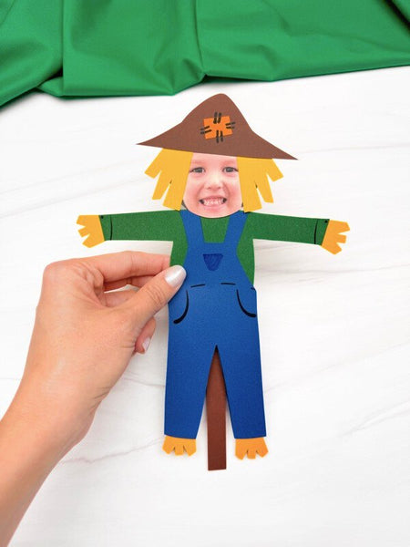 hand holding scarecrow photo craft on white marble with green fabric on top