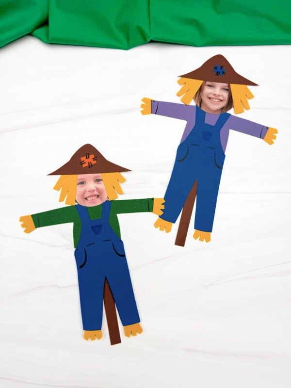 Two children's scarecrow cutout figures with printed faces, wearing blue overalls and green or purple shirts, displayed on a white surface with a green fabric backdrop.
