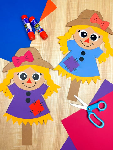 Girl scarecrow craft project with construction paper, glue, and scissors on a wooden surface.