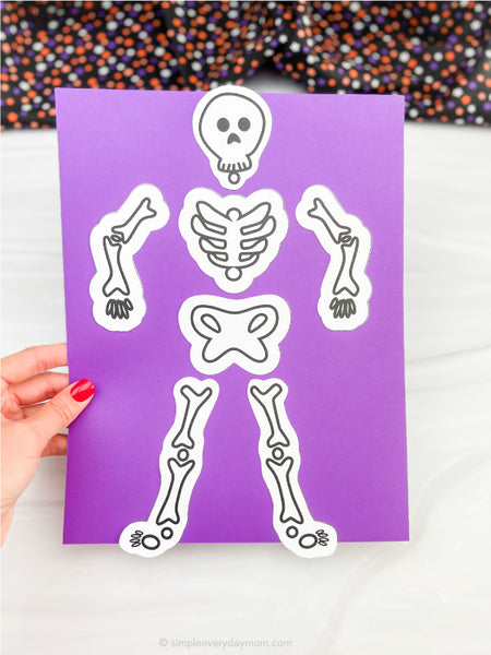 A hand with red nail polish holds a purple craft sheet featuring cut-out skeleton bone pieces with black outlines and white borders, set against a white surface with a colorful patterned fabric in the background.