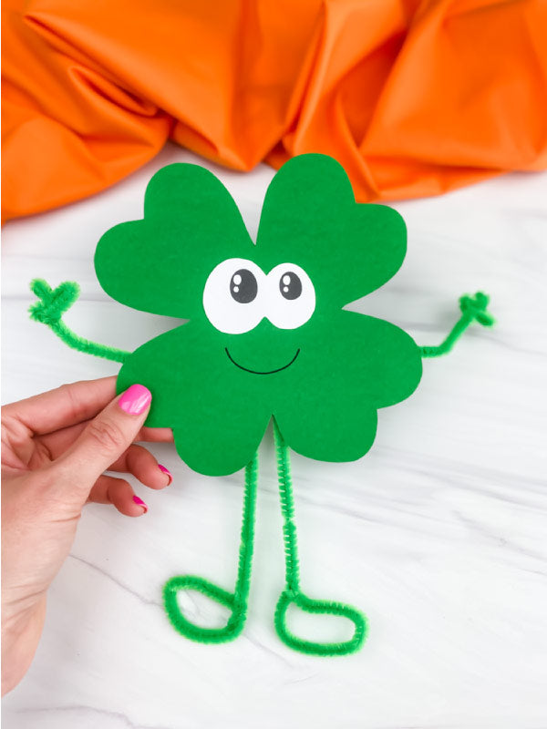 Handmade green paper shamrock with googly eyes and pipe cleaner arms and legs against an orange and white background.