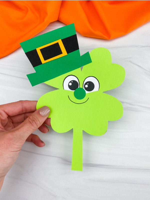 Hand holding a green paper shamrock with a leprechaun hat against an orange fabric background