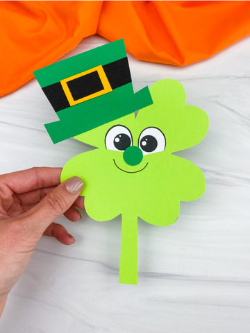 Hand holding a green paper shamrock with a leprechaun hat against an orange fabric background