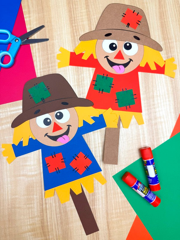 Two paper scarecrow crafts with colorful clothing on a wooden surface, surrounded by craft supplies.