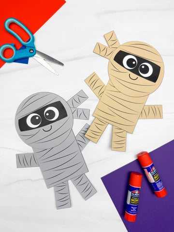 Two paper mummy crafts with scissors and glue sticks on a colorful background.