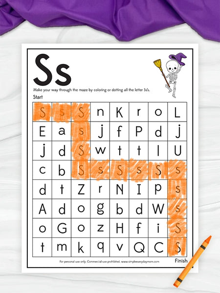 A printable letter S word maze worksheet with a grid of lowercase letters, decorative header text, a small cartoon skeleton, and orange highlight marks over several letters. The sheet is photographed on a white marble-like surface with purple fabric at the top and an orange pencil at the bottom right.