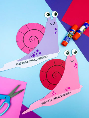 Two Valentine snail crafts on blue background with scissors, paper, and glue sticks. 